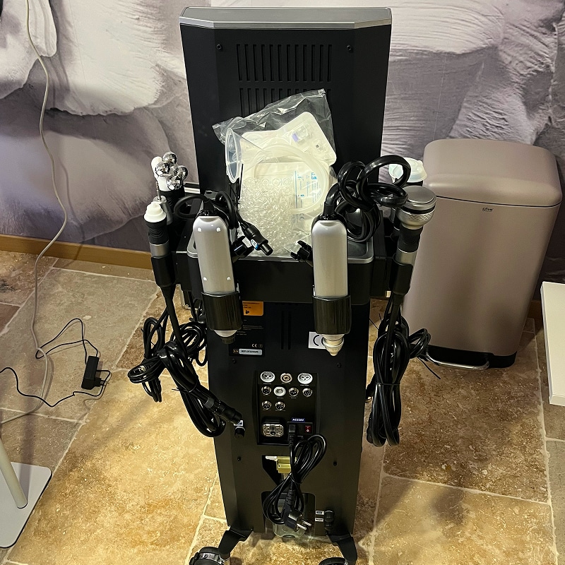 Arrière d'une machine de beauté multifonction noire sur roulettes, avec plusieurs poignées et connecteurs, et un sachet de consommables.