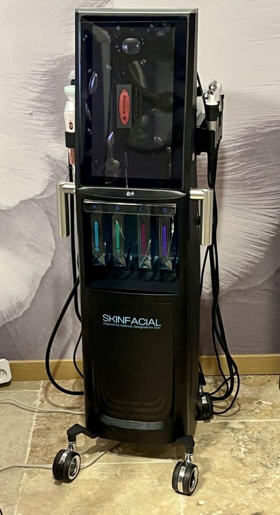 Machine SKINFACIAL : Traitement de la peau professionnel sur roulettes Appareil professionnel SKINFACIAL noir sur roulettes, avec quatre réservoirs de liquides colorés (A, B, C, D) et un avertissement rouge sur l'écran supérieur.